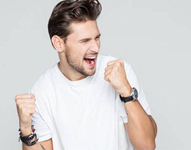 A man in a white t-shirt is celebrating enthusiastically with clenched fists, his eyes closed, and his mouth open.